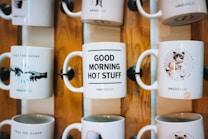 Several ceramic mugs are displayed on a wooden rack. Each mug features a different design and message. One mug reads 'PASS THE SUGAR' with an image of hands extended toward each other, another says 'GOOD MORNING HOT STUFF,' and a third features a grumpy cat with the message 'NOT TALKING TILL I GET MY COFFEE.' The mugs are arranged in two rows, showcasing a decorative and fun style.