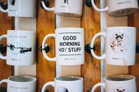 Several ceramic mugs are displayed on a wooden rack. Each mug features a different design and message. One mug reads 'PASS THE SUGAR' with an image of hands extended toward each other, another says 'GOOD MORNING HOT STUFF,' and a third features a grumpy cat with the message 'NOT TALKING TILL I GET MY COFFEE.' The mugs are arranged in two rows, showcasing a decorative and fun style.