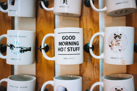 Several ceramic mugs are displayed on a wooden rack. Each mug features a different design and message. One mug reads 'PASS THE SUGAR' with an image of hands extended toward each other, another says 'GOOD MORNING HOT STUFF,' and a third features a grumpy cat with the message 'NOT TALKING TILL I GET MY COFFEE.' The mugs are arranged in two rows, showcasing a decorative and fun style.
