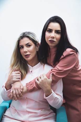 Two women are seated and closely embracing. The woman on the left has long blonde hair and is wearing a light pink blouse, while the woman on the right has dark hair and is leaning into the first woman, wearing a darker pink outfit. Their expressions appear calm and thoughtful.