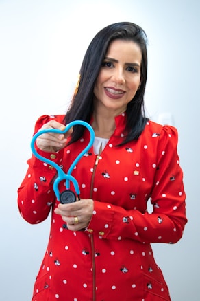 A woman with dark hair, wearing a red jacket adorned with white polka dots and small cartoon characters, is holding a stethoscope shaped in a heart. She is smiling and appears to be in a well-lit, indoor setting with a neutral background.