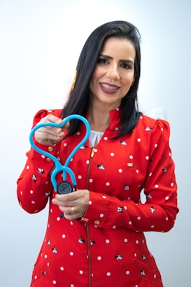 A woman with dark hair, wearing a red jacket adorned with white polka dots and small cartoon characters, is holding a stethoscope shaped in a heart. She is smiling and appears to be in a well-lit, indoor setting with a neutral background.