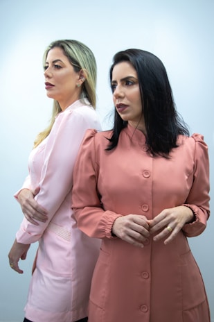 Two women are standing side by side against a light background. They are wearing outfits in shades of pink and peach, and their expressions are focused and serious.