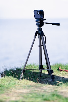 A camera mounted on a tripod is situated on a grassy surface with the background showing a blurred view, possibly of water. The tripod appears sturdy with adjustable legs and is positioned on uneven terrain, capturing a serene and natural setting.