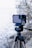 A smartphone is mounted on a tripod, equipped with an external lens attachment, in an outdoor setting. The background features a blurred view of a lake or sea, with bare trees and rocks, indicating a natural scene.