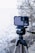 A smartphone is mounted on a tripod, equipped with an external lens attachment, in an outdoor setting. The background features a blurred view of a lake or sea, with bare trees and rocks, indicating a natural scene.