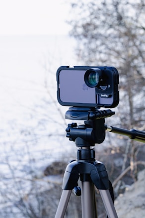 A smartphone is mounted on a tripod, equipped with an external lens attachment, in an outdoor setting. The background features a blurred view of a lake or sea, with bare trees and rocks, indicating a natural scene.