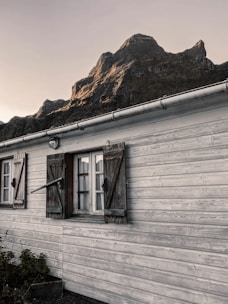 A rustic wooden house with shuttered windows stands in the foreground, characterized by weathered wood and simple design. In the background, a dramatic mountain rises under the soft lighting of either sunrise or sunset, casting shadows and highlighting its rugged contours.