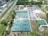 An aerial view of a sports complex featuring multiple tennis courts and a basketball court, surrounded by trees and a parking area with vehicles. There are paths leading to the courts, and a few small buildings nearby.