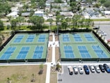 Multiple tennis courts are visible, each with blue and green playing surfaces. People are playing tennis, and the courts are surrounded by a tall fence. The courts are located near a parking lot with several parked cars. Trees and residential houses are in the background, creating a suburban setting.