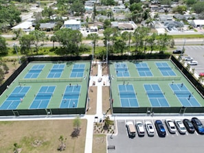 Multiple tennis courts are visible, each with blue and green playing surfaces. People are playing tennis, and the courts are surrounded by a tall fence. The courts are located near a parking lot with several parked cars. Trees and residential houses are in the background, creating a suburban setting.