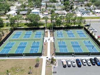 Multiple tennis courts are visible, each with blue and green playing surfaces. People are playing tennis, and the courts are surrounded by a tall fence. The courts are located near a parking lot with several parked cars. Trees and residential houses are in the background, creating a suburban setting.