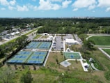 Aerial view of a recreational area featuring multiple tennis courts with blue surfaces, a basketball court, and a playground with slides. The facility is surrounded by greenery and a parking area with numerous cars. In the distance, a city skyline is visible on a clear day with scattered clouds.