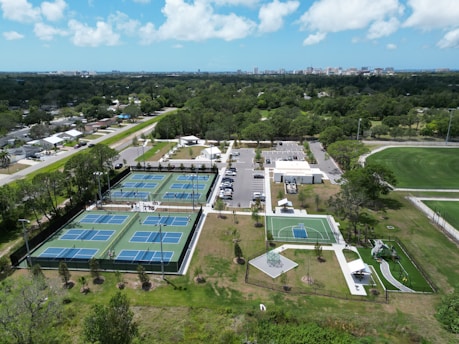 Aerial view of a recreational area featuring multiple tennis courts with blue surfaces, a basketball court, and a playground with slides. The facility is surrounded by greenery and a parking area with numerous cars. In the distance, a city skyline is visible on a clear day with scattered clouds.