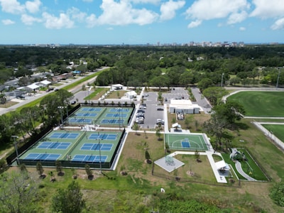 Aerial view of a recreational area featuring multiple tennis courts with blue surfaces, a basketball court, and a playground with slides. The facility is surrounded by greenery and a parking area with numerous cars. In the distance, a city skyline is visible on a clear day with scattered clouds.