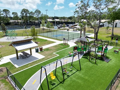 The image depicts a vibrant outdoor playground area featuring a playground set with swings, slides, and climbing structures on green turf. Nearby is a covered picnic area with a table and benches. In the background, there are tennis or basketball courts enclosed by fences, with trees and some parked cars visible. Bright natural lighting suggests a sunny day.