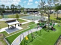 The image depicts a vibrant outdoor playground area featuring a playground set with swings, slides, and climbing structures on green turf. Nearby is a covered picnic area with a table and benches. In the background, there are tennis or basketball courts enclosed by fences, with trees and some parked cars visible. Bright natural lighting suggests a sunny day.