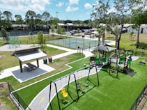 The image depicts a vibrant outdoor playground area featuring a playground set with swings, slides, and climbing structures on green turf. Nearby is a covered picnic area with a table and benches. In the background, there are tennis or basketball courts enclosed by fences, with trees and some parked cars visible. Bright natural lighting suggests a sunny day.