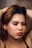 A person with closed eyes, wearing sparkly eye makeup and bold lipstick, poses with hands positioned behind the head. The backdrop is a textured, glittery surface, complementing the makeup's glamour.