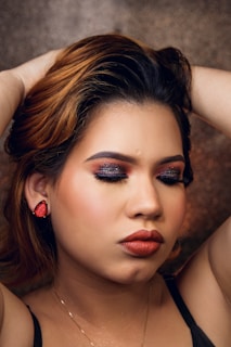 A person with closed eyes, wearing sparkly eye makeup and bold lipstick, poses with hands positioned behind the head. The backdrop is a textured, glittery surface, complementing the makeup's glamour.