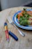 A wooden table is set with a plate of colorful food, including leafy greens, orange carrots, red pomegranate seeds, and broccoli, alongside a fork. Next to it, there is a blue and red insulin pen with a removable cap.