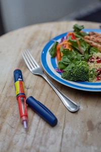 A wooden table is set with a plate of colorful food, including leafy greens, orange carrots, red pomegranate seeds, and broccoli, alongside a fork. Next to it, there is a blue and red insulin pen with a removable cap.