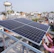 Two people are installing a large solar panel on a rooftop in an urban area with numerous buildings and a water tower in the background. The scene suggests an effort to set up renewable energy resources.