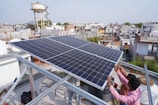 Two people are installing a large solar panel on a rooftop in an urban area with numerous buildings and a water tower in the background. The scene suggests an effort to set up renewable energy resources.