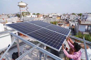 Two people are installing a large solar panel on a rooftop in an urban area with numerous buildings and a water tower in the background. The scene suggests an effort to set up renewable energy resources.