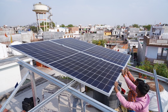 Two people are installing a large solar panel on a rooftop in an urban area with numerous buildings and a water tower in the background. The scene suggests an effort to set up renewable energy resources.