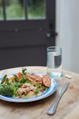A wooden table hosts a colorful meal featuring grilled chicken, couscous, green vegetables like broccoli and spinach, and red bell peppers on a blue-bordered white plate. A glass of water and a couple of pills sit nearby, alongside a fork and knife.
