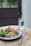 A wooden table hosts a colorful meal featuring grilled chicken, couscous, green vegetables like broccoli and spinach, and red bell peppers on a blue-bordered white plate. A glass of water and a couple of pills sit nearby, alongside a fork and knife.