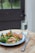 A wooden table hosts a colorful meal featuring grilled chicken, couscous, green vegetables like broccoli and spinach, and red bell peppers on a blue-bordered white plate. A glass of water and a couple of pills sit nearby, alongside a fork and knife.