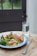 A wooden table hosts a colorful meal featuring grilled chicken, couscous, green vegetables like broccoli and spinach, and red bell peppers on a blue-bordered white plate. A glass of water and a couple of pills sit nearby, alongside a fork and knife.