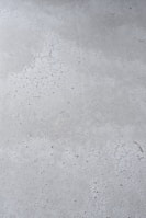 A close-up view of a concrete surface with visible cracks and a slightly textured appearance. The surface is predominantly gray with some darker specks scattered throughout, adding to the rough texture.