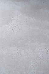 A close-up view of a concrete surface with visible cracks and a slightly textured appearance. The surface is predominantly gray with some darker specks scattered throughout, adding to the rough texture.