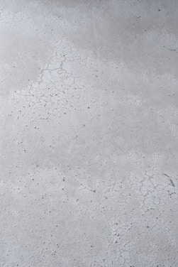 A close-up view of a concrete surface with visible cracks and a slightly textured appearance. The surface is predominantly gray with some darker specks scattered throughout, adding to the rough texture.