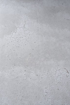 A close-up view of a concrete surface with visible cracks and a slightly textured appearance. The surface is predominantly gray with some darker specks scattered throughout, adding to the rough texture.