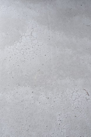 A close-up view of a concrete surface with visible cracks and a slightly textured appearance. The surface is predominantly gray with some darker specks scattered throughout, adding to the rough texture.