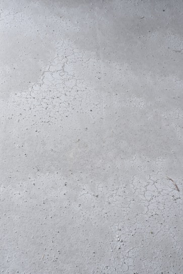 A close-up view of a concrete surface with visible cracks and a slightly textured appearance. The surface is predominantly gray with some darker specks scattered throughout, adding to the rough texture.