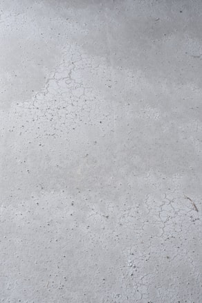 A close-up view of a concrete surface with visible cracks and a slightly textured appearance. The surface is predominantly gray with some darker specks scattered throughout, adding to the rough texture.
