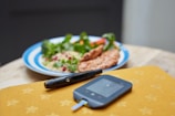 A glucometer and lancet are placed on a yellow surface with star patterns, while in the background, there is a plate of food with breaded items, couscous, and green vegetables.
