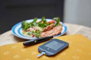 A glucometer and lancet are placed on a yellow surface with star patterns, while in the background, there is a plate of food with breaded items, couscous, and green vegetables.