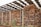 Stacks of cut logs are neatly arranged under a large metal roof. The logs are of various sizes and are piled high, enclosed by a chain-link fence. The structure has several metal posts supporting the roof, and there are small patches of grass and plants visible at the base.