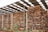Stacks of cut logs are neatly arranged under a large metal roof. The logs are of various sizes and are piled high, enclosed by a chain-link fence. The structure has several metal posts supporting the roof, and there are small patches of grass and plants visible at the base.