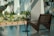 A modern interior space with large windows showcasing a lush green garden outside. The room features a minimalist design with a sleek brown lounge chair beside a small, round side table. Sunlight casts soft shadows across the polished concrete floor, enhancing the tranquil ambiance.