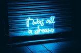 Neon blue text reading 'it was all a dream' is illuminated against a dark backdrop with metal shutters and tiled floor visible.