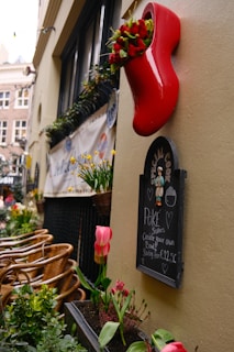 A cozy outdoor cafe scene with wooden chairs neatly arranged along a narrow pathway, surrounded by vibrant green plants and blooming tulips. A bright red decorative shoe mounted on the wall serves as a flower pot. Below the shoe, a chalkboard offers a special menu of poké bowls starting from €12.50.