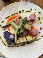 A beautifully plated dish featuring grilled fish, purple potatoes, radishes, asparagus, and a vibrant red sauce, garnished with green pesto drizzle.
