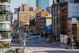 A city street scene features a mixture of modern and older brick buildings. There are large signs, including one advertising retail space for lease. The street is wide and relatively empty, with a few vehicles visible, including a yellow taxi. Architecture includes fire escapes and various styles of windows, with some greenery along the sidewalks.